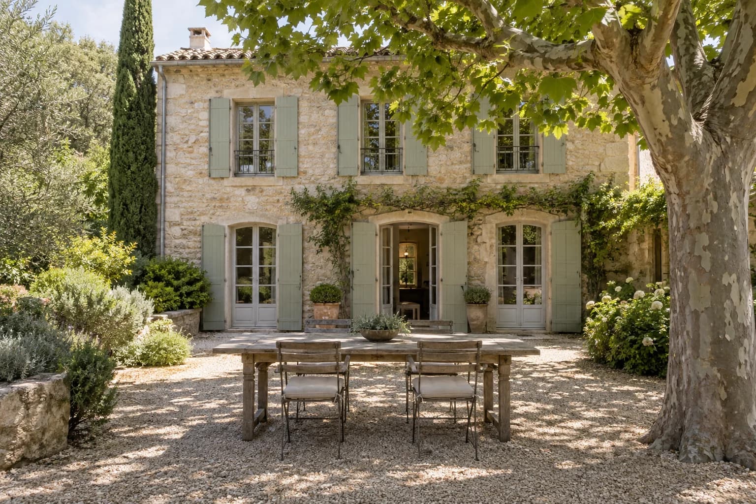 A stone family home with green shutters and a dining table beneath a plane tree.